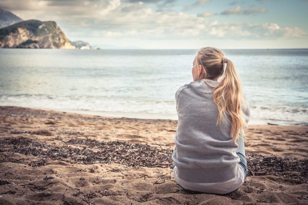 Pensive lonely young woman sitting on beach hugging her knees and looking into the distance with hope